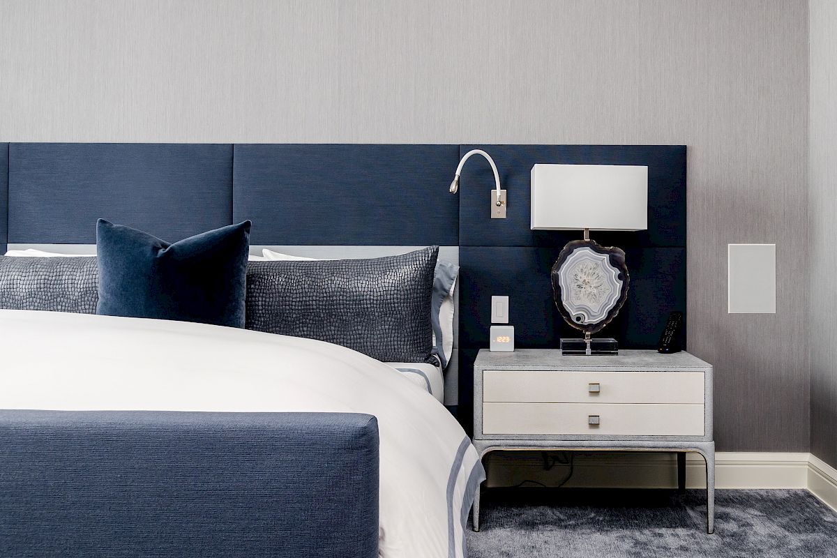 A modern bedroom with a blue and white color scheme, featuring a bed with dark cushions, a bedside table, and a lamp next to it.