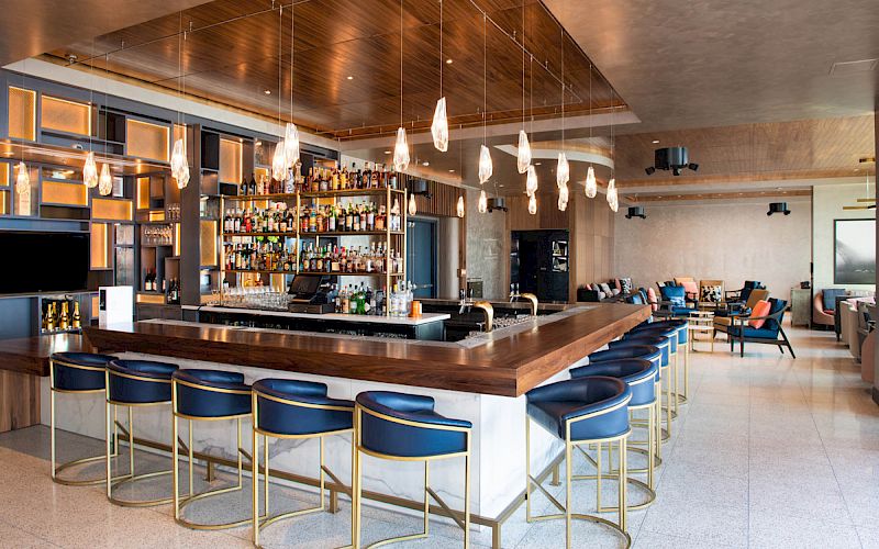 The image shows a modern bar with blue barstools, a well-stocked bar area, hanging lights, and a seating area with tables and chairs in the background.