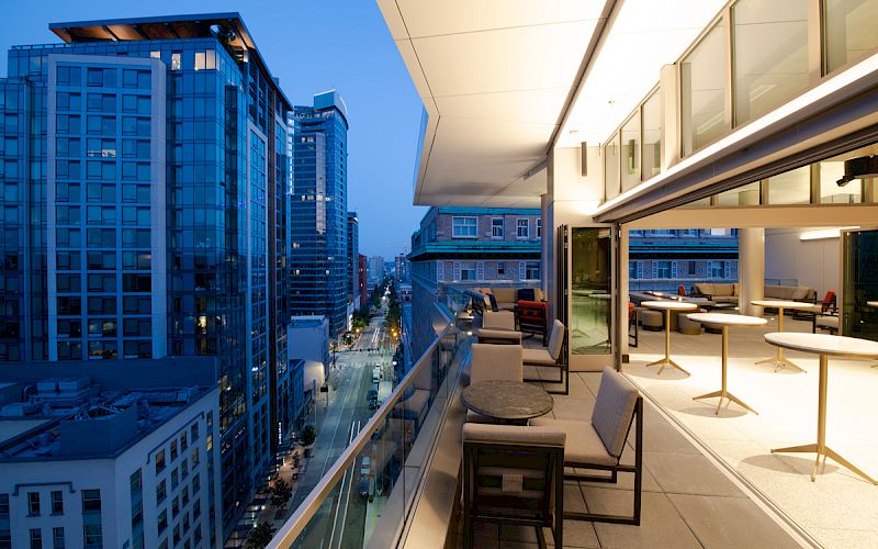 A modern cityscape is seen from a high-rise balcony at twilight, featuring outdoor seating and a view of glass-covered buildings lining a street.