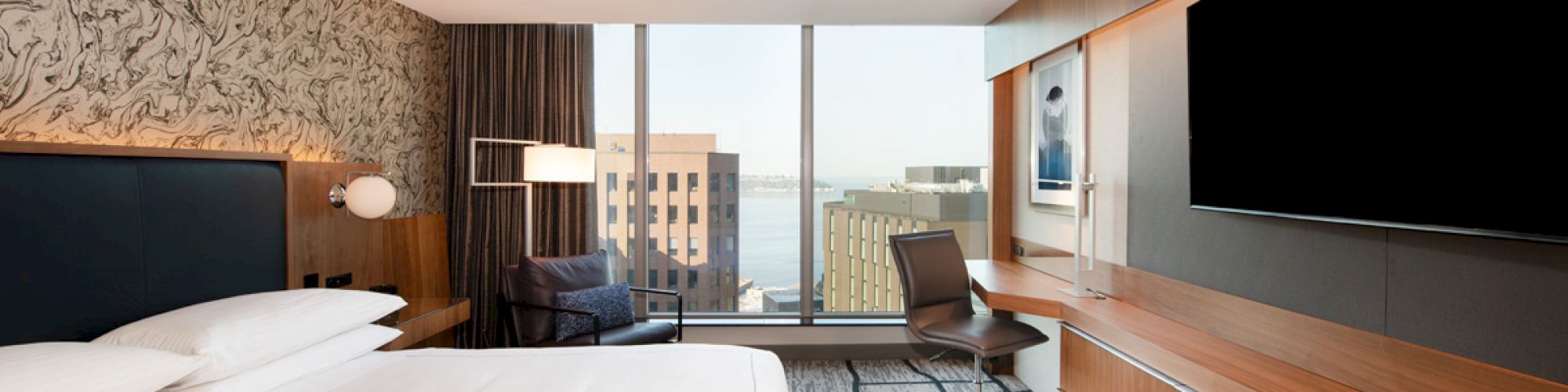 A modern hotel room featuring a large bed, wall TV, desk, and chair, with a view of buildings and water through a large window.