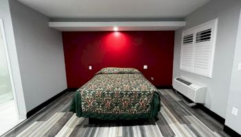 A simple bedroom with a double bed, floral bedspread, red accent wall, grey flooring, a window with white shutters, and a wall-mounted AC unit.