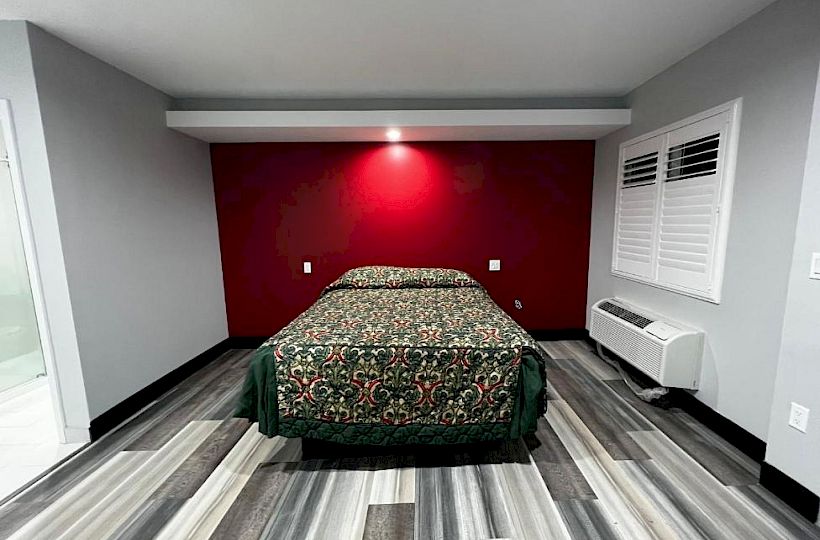 A minimalist room with a bed against a red accent wall, patterned bedspread, grey flooring, a window with blinds, and an air conditioning unit.