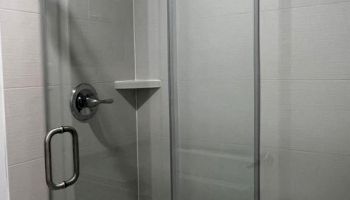 The image shows a modern glass shower enclosure with sliding doors, a handle, and a built-in shelf. The wall tiles are light-colored.