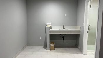 The image shows a clean, grey-tiled room with a sink and countertop, a small trash can underneath, and an adjacent door leading to a shower area.