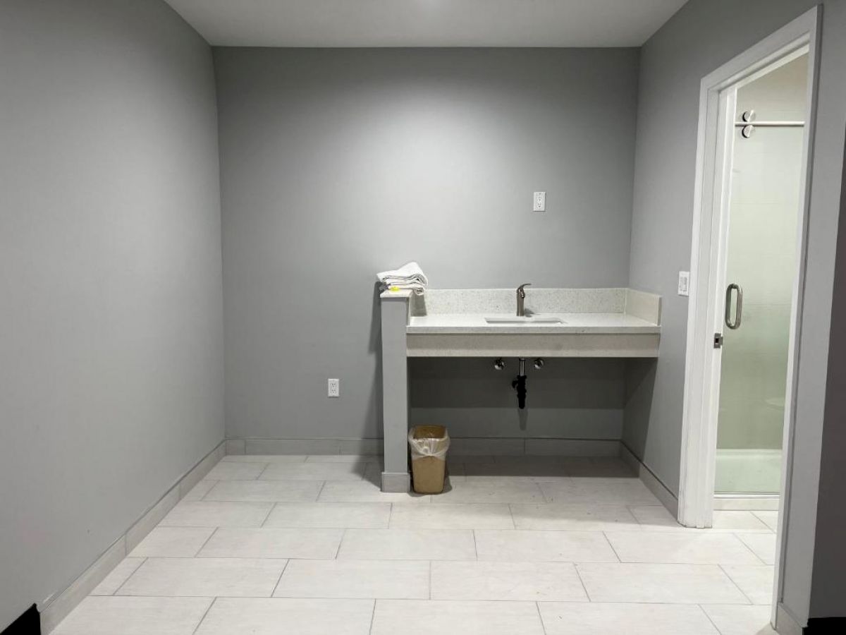 The image shows a room with a sink, garbage bin, some folded towels, and a door leading to another room or possibly a shower.