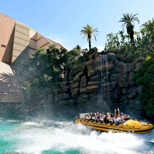 A group of people on a raft enjoying a water ride with a splash under a waterfall in a theme park, surrounded by tropical greenery and a large building.