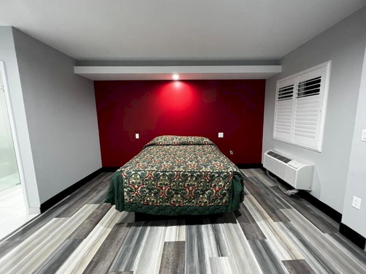 A modern bedroom with a patterned bedspread, red accent wall, grey flooring, and white walls with a window and air conditioning unit.