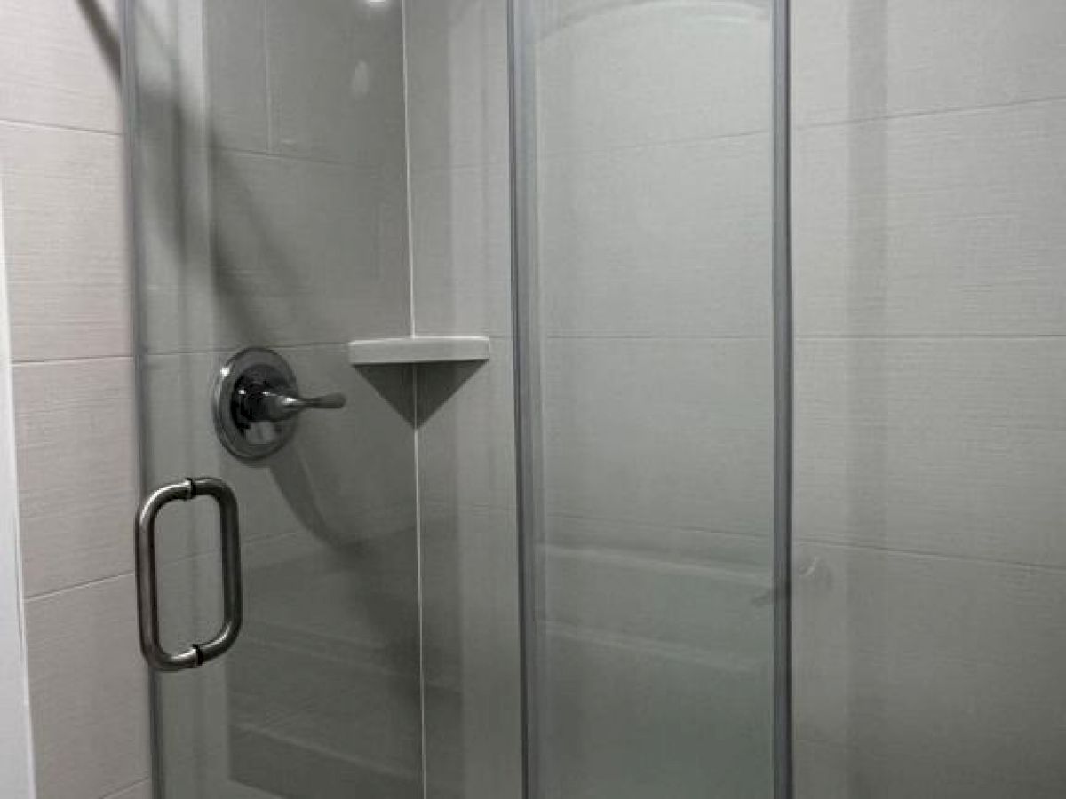 A modern shower with a glass door, chrome handle, and wall-mounted showerhead, has a built-in corner shelf against tiled walls.